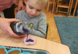 Chłopiec z pomocą nauczyciela napełnia powstałym fioletowym roztworem silikonową foremkę w kształcie serca.